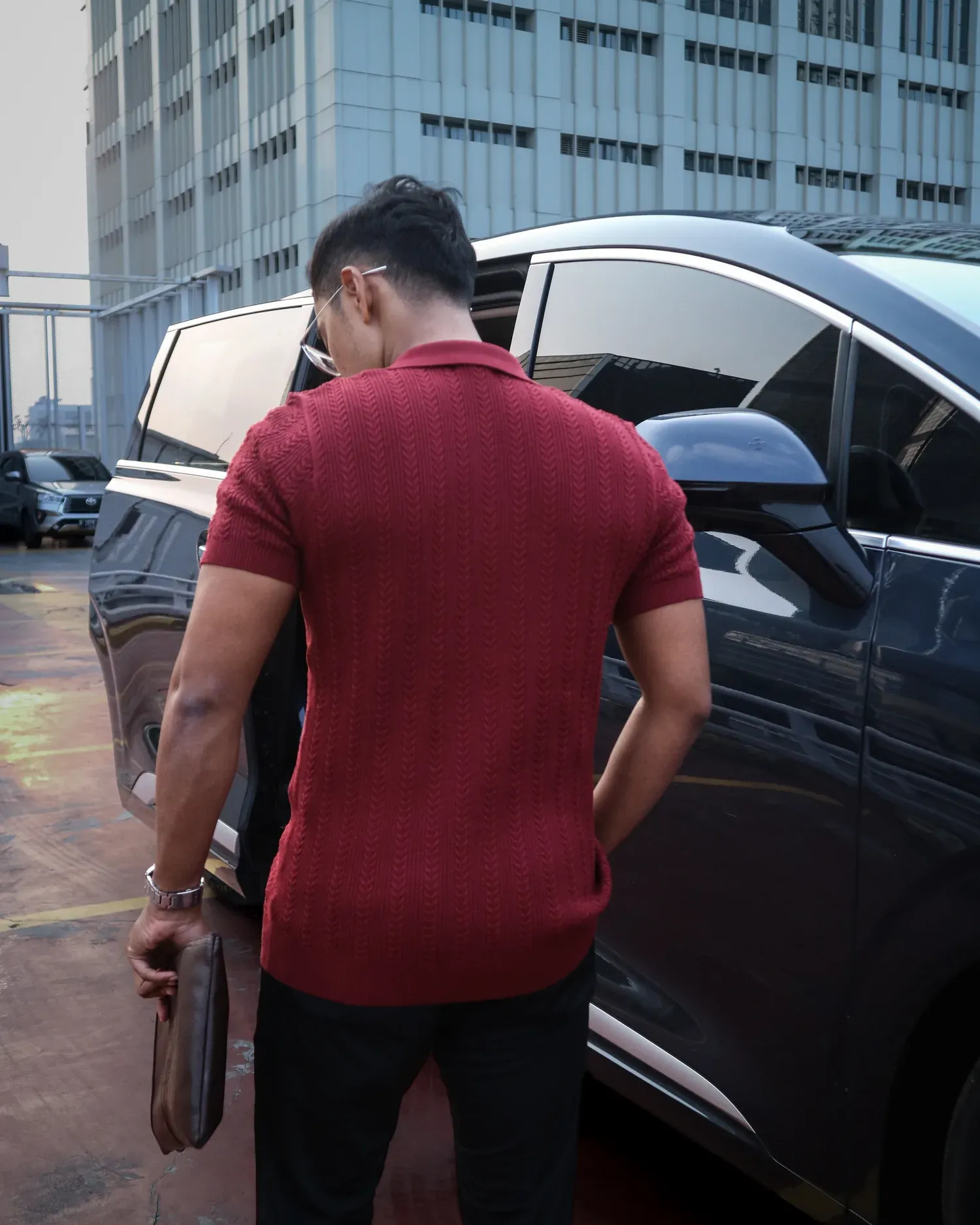 Tampilan belakang Mazly Signature Polo marun muda saat berdiri di dekat mobil
