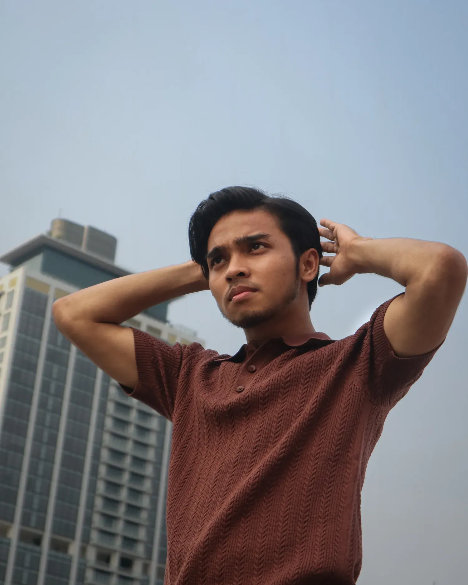 Pria mengenakan Mazly Signature Polo cokelat tua dengan kedua tangan di belakang kepala, berlatar gedung kota